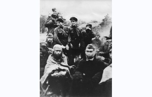 Grupa jeńców sowieckich grzejących się przy zapalonym ognisku. Niemiecki wartownik uzbrojony w karabin w płaszczu i furażerce.